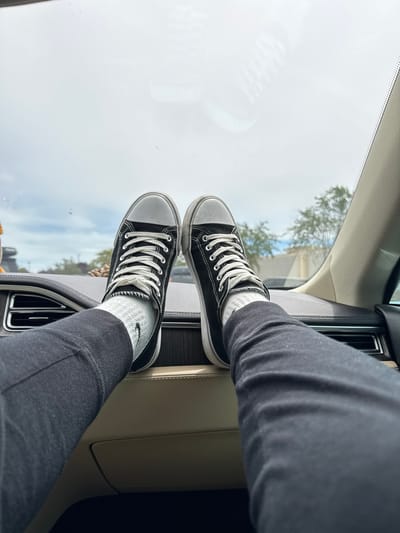 In Lakewood, United States of America, a close-up, first-person perspective shot captures a pair of feet resting on a car's dashboard. The individual's legs, clad in black pants, extend upwards, with feet adorned in black canvas sneakers and white socks. The sneakers have white laces and rubber soles. The car's interior, featuring a dark dashboard with horizontal vents and a beige trim, is visible. Beyond the windshield, a cloudy sky and distant trees can be seen. The scene is static, with no apparent interaction or action.