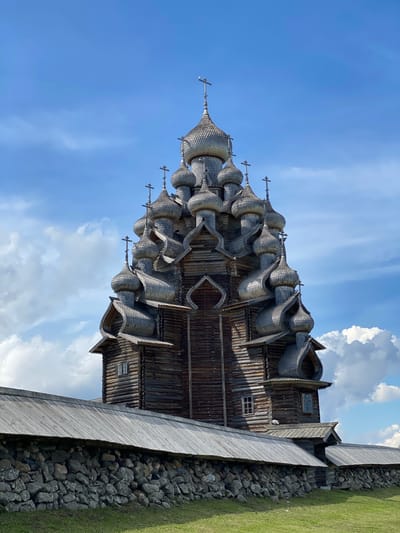 The image shows the Kizhi Pogost, a historical architectural ensemble located on Kizhi Island in the Republic of Karelia, Russia, near Boyarshchina. The dominant structure is a large, elaborately constructed wooden church with numerous onion domes, each topped with a cross. The church is made of dark wood logs and features intricate architectural details, including curved rooflines and decorative elements. In the foreground, a low stone wall with a wooden roof runs horizontally across the frame, separating the church from a grassy area. The sky above is blue with scattered white clouds. The scene is captured from a medium distance, with the church being the primary subject. There are no people or animals visible in the image.