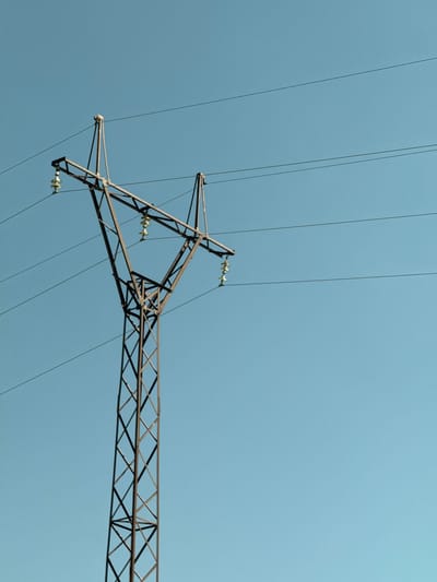 An overhead power line tower stands against a clear, light blue sky. The tower is constructed from dark metal lattice work, forming a tall, tapering structure. At the top, it branches out to support multiple high-voltage electrical cables, which stretch horizontally across the frame. Several insulators, appearing yellowish-white, are visible along the cables, attached to the tower's crossbars. The image is taken from a low angle, emphasizing the height of the tower and its overhead infrastructure. The scene is devoid of any people, animals, or other structures. This infrastructure is located in Tsovagyugh, Armenia.