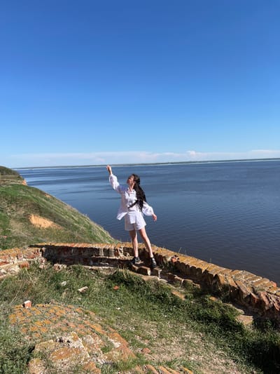 A medium shot angled with a woman in a white outfit standing on a wall by a body of water under a blue sky.
The woman stands on a crumbling brick wall with vegetation growing on it. She wears a long-sleeved white top and white shorts, with dark footwear. Her dark hair flows down her back. She has one arm outstretched, pointing towards the water. The water stretches to the horizon, where a thin line of green land is visible. The sky is a clear, light blue.
Based on geolocation, the location is near Шуран, Russia.
