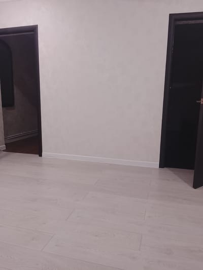 The image shows an interior space with light wood-look flooring. Two dark-framed doorways are visible on either side of a light-colored wall. The doorway on the left appears to lead into a room with darker walls and trim. The doorway on the right is partially open, revealing a dark interior. The location is given as Assinovskaya, Russia. There are no people visible.