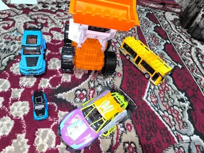 The scene is an indoor setting within the city of Pakhtakor, Uzbekistan, viewed from a slightly elevated perspective. Five toy vehicles are arranged on a dark red and gray patterned carpet. No individuals are present.
In the upper center, a large orange and white construction vehicle, resembling a front-end loader, is positioned with its rear facing the camera, displaying black wheels and an elevated orange bucket. To its immediate left, a medium-sized blue SUV-style toy car features a black hood detail and a transparent roof section. Below this blue SUV, a smaller blue and black toy car rests. To the right of the large construction vehicle, a yellow double-decker bus toy lies horizontally, displaying multiple windows. In the lower center, a purple and yellow futuristic-style race car toy with prominent spoilers and exposed wheels is present. A dark, possibly curtained, fabric with an indistinct pattern is partially visible in the upper right background, suggesting room furnishing.