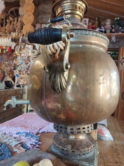 This image captures the interior of a rustic establishment, likely a shop or cafe, located in Staraya Russa, Russia. The central focus is a large, ornate, tarnished brass samovar positioned on a wooden table in the foreground.

The samovar features a reflective, bulbous body showing signs of age and patina. Its handle, crafted from dark, possibly wooden, material with brass fittings, is wrapped with a light, coiled cord. A decorative brass spigot with an animal-head motif extends from the lower front of the samovar. The samovar rests on an intricate brass base with perforated rectangular designs.

To the left of the samovar on the wooden table, a multi-textured, hand-woven or crocheted textile, presenting shades of pink, blue, and brown, is spread. In the bottom left corner, a dark, shallow bowl is partially visible, containing a single green, spherical fruit and several small, white, spherical objects.

The background reveals a rustic setting with prominent wooden elements, including walls and a sloped ceiling. On the left side, shelves are densely packed with various small items, suggesting souvenirs or local crafts, while bundles of dried goods hang above. A large wooden wagon wheel is mounted on the upper left wall. Further back on the left, behind a counter, a person with light-colored hair is visible, appearing to be an attendant, operating a point-of-sale system. On the right side of the background, additional shelves display an assortment of soft toys and decorative items, including a small pink doll and a grey stuffed frog. A black-on-white arrow sign is partially visible on the far right.