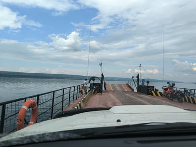 The image depicts a ferry crossing a body of water, likely the Angara River, in Boguchany, Russia. The foreground shows the hood and windshield of a vehicle, indicating the viewpoint is from inside a car waiting to disembark. The ferry itself is a utilitarian vessel with a wide deck designed to transport vehicles.
On the ferry's deck, several people are visible. One man in a white shirt and shorts stands near a railing, looking out at the water. Another person, wearing a dark cap, is interacting with a motorcycle. Two motorcycles are parked on the ferry: one orange, and another partially obscured green bicycle. There are also yellow and black striped markings on the deck, likely indicating safety zones or vehicle parking spots.
A bright orange life preserver with white stripes is attached to the railing on the left side of the ferry. In the background, a Russian flag flutters on a tall pole, and another, smaller flag with red, white, and blue colors is also visible. The water is calm and reflects the cloudy sky. Rolling hills or mountains form the distant horizon. The overall scene suggests a typical day on a river ferry, likely a common mode of transport in this region.