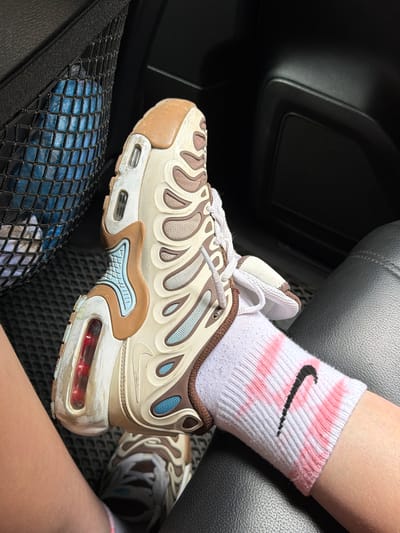 A close-up shot focuses on a person's legs and feet inside a vehicle, as indicated by the dark interior and car seat texture. The primary subject is a pair of beige and brown Nike Air Max Plus sneakers, worn with white crew socks that have a pink tie-dye pattern and the iconic black Nike swoosh logo. The shoes appear well-worn, with some scuff marks and dirt visible on the soles and white sections. The person's legs are bare from the knee down. In the foreground, a black mesh pocket is visible, containing a blue object, possibly a water bottle. The lighting suggests it might be daytime, possibly with natural light filtering into the car. The image seems to capture a casual moment, with no strong emotions conveyed. The setting appears to be inside a car, possibly on a trip given the inclusion of a mesh pocket often found in vehicles. The location context provided is Turbaza Katun', Russia, but this is not directly discernible from the visual elements of the image itself.