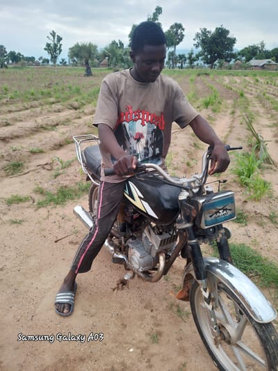 A young Black male sits on a motorcycle in a rural area. He is wearing a light brown t-shirt with a graphic design on it, dark pants with pink stripes, and grey flip-flops. The motorcycle is black and silver with a chrome exhaust pipe and handlebars. The background shows a field with rows of plants, trees, and huts, suggesting a village setting in Angwan Hausawa, Nigeria. The sky is overcast. The Samsung Galaxy A03 watermark is visible in the bottom left corner of the image.