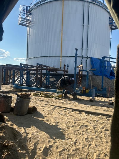 A worker in a grey jumpsuit and white hard hat is welding in front of a large white cylindrical tank in Чикшино, Russia. The tank has a grated walkway and ladder on its left side and green pipes running vertically along its surface. A blue tarp is draped over some construction materials to the right of the tank. The ground is covered in sand with numerous footprints. Several buckets, gloves, and rolled-up materials are scattered on the sand in the foreground. The scene appears to be a construction or industrial site under a clear blue sky with a few clouds.
