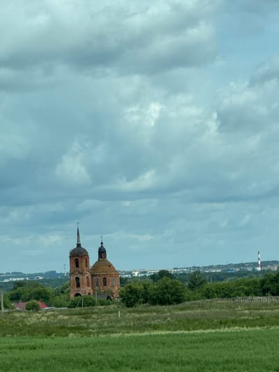 The image captures a landscape scene with an old, abandoned brick church as the central focus. The church has two domes, one taller and more slender with a spire, and another shorter and rounder. Both domes are topped with crosses. The brickwork of the church is weathered and appears to be in a state of disrepair.
The church is situated behind a field of green grass and vegetation. In the mid-ground, there are dense trees surrounding the church and a few scattered buildings with red roofs. Further in the background, a city skyline is visible, characterized by various buildings and a tall, red and white striped industrial chimney on the right side of the frame. The sky is filled with heavy, grey clouds, suggesting an overcast or impending stormy weather. The overall impression is one of a rural setting with a historical religious structure set against a backdrop of industrial development.
The image is taken from a slightly elevated position, looking across the field towards the church and the distant city. No people or animals are visible in the frame. The lighting is diffused due to the cloud cover. The scene is static, with no apparent action occurring. The location is identified as Tatarstanskiye Chelny, Russia.