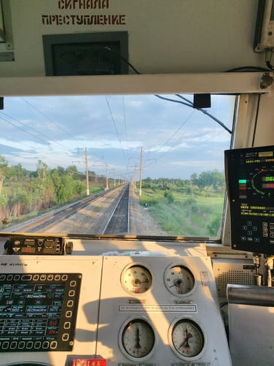 This media file shows the interior view from the cab of a train, looking out at the tracks stretching into the distance. The scene appears to be set in Bureya, Russia. The weather is fair with a partly cloudy sky, and the time of day seems to be late afternoon or early evening, indicated by the warm, golden light illuminating the cab's controls.

The primary subjects are the various control panels and instruments within the train's cab. These include a display screen showing operational data like speed, distance, and time, as well as analog gauges for pressure and other indicators. Prominent text visible on the panels includes "СИГНАЛА ПРЕСТУПЛЕНИЕ" (Signal of Crime/Violation) in red stencil letters on the ceiling, and "НЕ КУРИТЬ" (No Smoking) and "НЕ ВСКРЫВАТЬ ПРИ ПОДНЯТОМ ТОКОПРИЕМНИКЕ" (Do Not Open When The Pantograph Is Raised) on smaller signs.

The setting is the driver's compartment of a train, with a wide window offering a view of the railway line ahead. The landscape outside is characterized by lush green trees and fields, with power lines running alongside the tracks, typical of a rural or semi-rural environment in Russia.

The activity is implied to be the operation of the train, with the driver presumably in control, as evidenced by the complex instrumentation. There are no people clearly visible in the image, but the perspective suggests someone is occupying the driver's seat. The overall mood is functional and operational, with the natural light creating a somewhat serene atmosphere despite the technical nature of the environment.