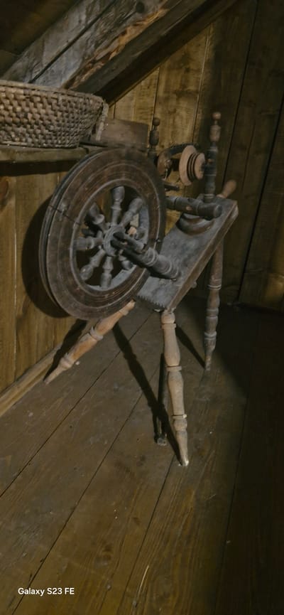 The image depicts a dimly lit attic or storage space, likely within the Pakruojis Manor in Lithuania, given the historical context of such artifacts. The foreground is dominated by a vintage wooden spinning wheel, a complex mechanism with a large spoked wheel on the left, a spindle assembly, and a wooden frame supported by turned legs. The wood appears aged and weathered. To the left and slightly behind the spinning wheel, a woven basket is visible, resting on a wooden surface. The background consists of wooden walls and a sloping wooden ceiling, suggesting an old building structure. The lighting creates strong shadows, emphasizing the textures of the wood and the aged appearance of the objects. There are no people or animals present in the scene. The overall atmosphere is one of quiet history and preservation.
