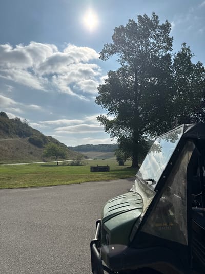 A partial view of a green utility terrain vehicle (UTV) with a windshield and a dark canvas soft top occupies the right foreground, positioned on an asphalt surface. The scene is located in Wold Newton, United Kingdom, specifically in a rural landscape.
The midground features an expansive green grassy field. A lone, small deciduous tree stands towards the left-center of this field. Further back, centrally located within the field, is a dark, rectangular, low-lying structure, appearing to be a raised planter or trough. Two large, mature deciduous trees with full green foliage are situated on the right side of the midground, partially obscured by the UTV.
The background on the left consists of a steep, treeless hill covered in light brown and green vegetation, indicative of dry conditions. A narrow, light-colored track or path is visible traversing the slope of this hill. The distant background on the right reveals more rolling hills and cultivated fields under a clear sky with scattered white, layered clouds. The sun is prominently visible in the upper center of the sky, casting a bright glare, suggesting a daytime scene. No individuals, actions, or specific interactions are depicted.