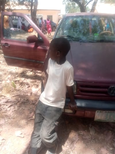 An African male child, approximately 5-7 years old, stands in the foreground, facing right with his head slightly turned. He wears a light-colored, short-sleeved t-shirt and dark trousers. His left hand rests on the front bumper of a maroon sedan, while his right side is positioned near the open driver's side door. The sedan displays a Volkswagen emblem on its front grille and a partially visible Nigerian license plate bearing "LAGOS" and "J J". In the mid-ground, several adults are observed near and around other vehicles. At least two males in white garments and two females, one in a vibrant pink dress and another less distinct, are present. The environment consists of unpaved, reddish-brown dirt ground with scattered dry leaves and sparse vegetation. Bright daylight casts shadows. Mature trees with sparse foliage and a light-colored building are visible in the background. The scene is located in Kagoma, Nigeria.