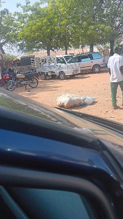 The image displays an outdoor scene in Langtang, Nigeria, under clear daytime conditions. The immediate foreground is obscured by the dark blue upper edge of a vehicle. The midground features a reddish-brown dirt surface where two large, light-colored sacks, one secured with a light blue strap, are resting. To the left, two motorcycles are parked, one with a visible red fuel tank. Adjacent to the motorcycles, a white utility truck with a caged bed containing multiple white packages or containers is parked. Behind the utility truck, a silver or light-colored SUV is also parked. On the far right, a male subject is observed from the rear, standing or walking, dressed in a white short-sleeved shirt, dark green trousers, and a dark green cap. The background consists of dense green trees and low-rise structures with corrugated metal roofs, under a bright, cloudless sky.