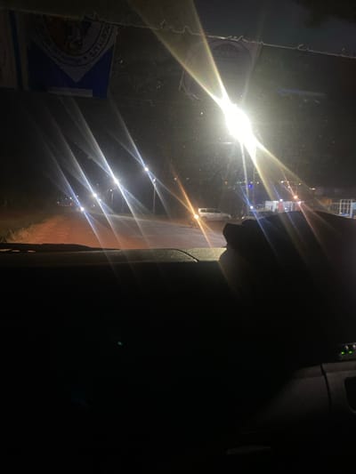 The image depicts a nighttime street scene in Kiaga, Kenya, observed from the interior of a vehicle. The lower portion of the frame shows the vehicle's dark dashboard, with the upper right section of a human hand visible resting on it. Through the windshield, a dirt or unpaved road extends into the distance, illuminated by multiple bright streetlights that create pronounced lens flares across the view. A white sport utility vehicle (SUV) is parked on the right side of the road in the mid-ground. Further down the road to the left, additional distant vehicle headlights are visible. In the background, beyond the parked SUV, indistinct structures with some external lighting indicate a populated area. The upper part of the windshield contains adhesive labels, one of which is partially legible as "SOCIETY LTD". A faint green light source is present on the far right, possibly from the vehicle's console.