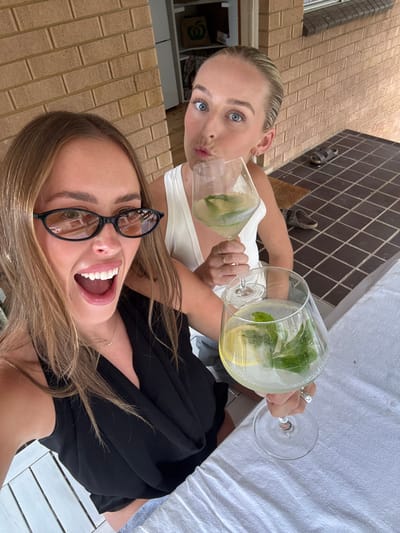 Two young women pose for a selfie, holding large glasses filled with a clear beverage, lemon slices, and mint leaves. The woman in the foreground smiles widely, wearing black-framed glasses and a black top. The woman behind her puckers her lips, wearing a white tank top. They appear to be outdoors on a patio, with a white tablecloth visible in the background. The setting includes brick walls and outdoor decor.