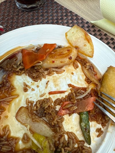 The image depicts a partially consumed meal on a white plate, likely in a restaurant or dining establishment in Lohfelden, Germany. The white, fluted plate contains remnants of a dish featuring brown rice, slices of orange-red bell pepper or carrot, white onion slices, and a green bean-like vegetable, all coated in a dark brown sauce and grease. A golden-brown, deep-fried item (possibly an egg roll) is visible on the right edge of the plate, and a golden-brown, flat, potato-like slice is at the top right. A silver four-tined fork is partially visible, resting on the right side of the plate. Above the plate, a dark beverage in a clear, faceted glass sits on a dark brown woven placemat. A light yellow or beige textured napkin is visible to the right of the placemat, on a light wood-grain table surface. No individuals are present in the frame.