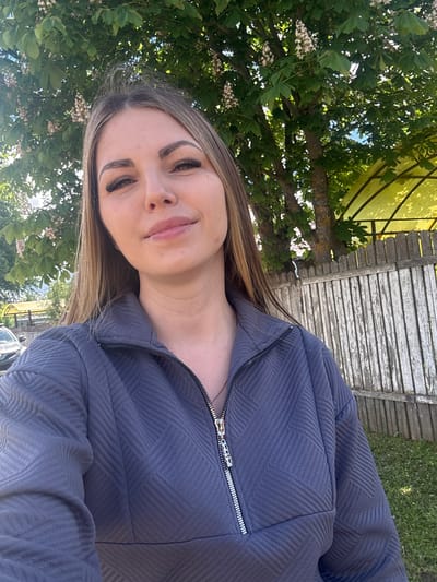 A woman with long blonde hair and false eyelashes is taking a selfie outdoors. She is wearing a blue textured zip-up jacket. Behind her is a tree with green leaves and white flowers, and a weathered wooden fence. The location is not identifiable as Slobodka, Russia from the information in the image.