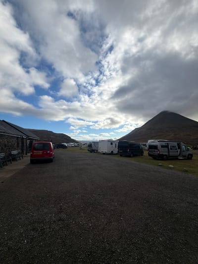 The image depicts a row of vans and campers parked along a gravel road in the Isle of Skye, United Kingdom. A red car is also parked nearby. The sky above features a mix of clouds and patches of blue. A mountain partially obscures the background. A building sits at the bottom edge of the image, next to the red car.