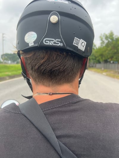 The image, taken from behind, shows a person wearing a black helmet and a black t-shirt, possibly riding a motorcycle. The helmet has several stickers, including a round white one with Vietnamese text and a QR code. The person has brown hair and is wearing a thin necklace. The background suggests an outdoor setting, likely on a road with trees and vegetation on the sides, consistent with a location in the city of Phúc Yên, Vietnam. The sky is overcast, and there are water droplets on the helmet, indicating it may have rained.
