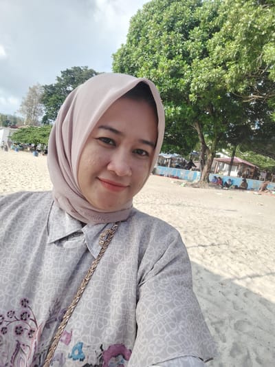 A medium shot captures a woman wearing a light pink hijab and a grey patterned shirt standing on a sandy beach. She is looking at the camera and smiling. Behind her, there is a large green tree, and further in the background, there are other trees, buildings, and people gathered. The sky is partly cloudy. The setting appears to be a beach in Parapat, Indonesia, with the distinct environment of a tropical location.