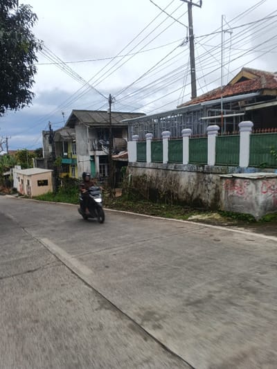A dark-colored scooter, operated by an individual wearing a dark shirt and helmet, travels on a gray paved road under an overcast sky. The specified location of Masaiti, Zambia, cannot be visually confirmed by the image.
On the right side of the road, a concrete retaining wall with green moss and red graffiti supports a fence featuring white pillars and green metal railings. Behind the fence, buildings include one with a reddish-brown tiled roof and exposed metal framing, suggesting construction or renovation, and another multi-story structure with a dark corrugated roof. On the left, further down the road, a small, light-brown building with an illegible sign and additional residential-style structures are visible. Multiple utility poles support a dense network of overhead power and communication lines that crisscross the sky. A dark tree canopy is present in the upper left quadrant. Sparse vegetation grows along the roadside, indicating an urban or semi-urban environment.