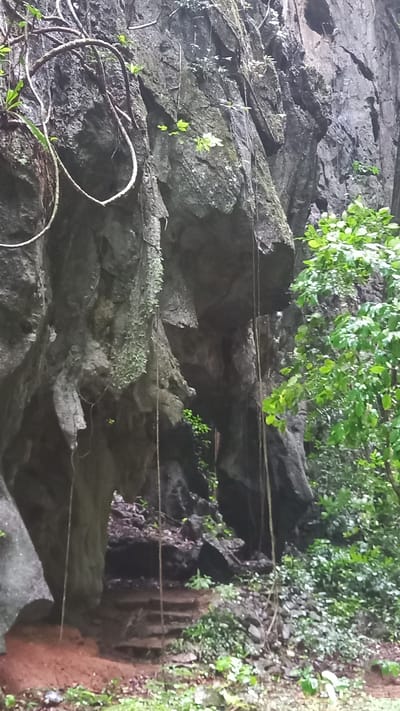 This is a photograph of a rock formation in Ban Pak Ra, Thailand. The rock is dark and heavily textured. There is greenery growing on and around the rocks, including bushes and vines. A length of rope runs across the scene horizontally, suspended at two points. To the right of the rocks are some steps that appear to be carved into the stone. There is also reddish soil visible on the right side of the image.