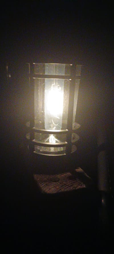 The image shows a close-up of an outdoor light fixture against a dark, possibly night, background. The light fixture is cylindrical, made of glass and metal. A bright light bulb is visible inside, casting a glow. Below the fixture, a rough-textured, dark-colored object, possibly wood, is visible. The overall scene is dark and grainy, suggesting low light conditions and possibly a low-resolution camera. The image does not provide any specific visual cues to determine its exact location within Lipí, Czechia, or to describe any people, actions, or interactions.