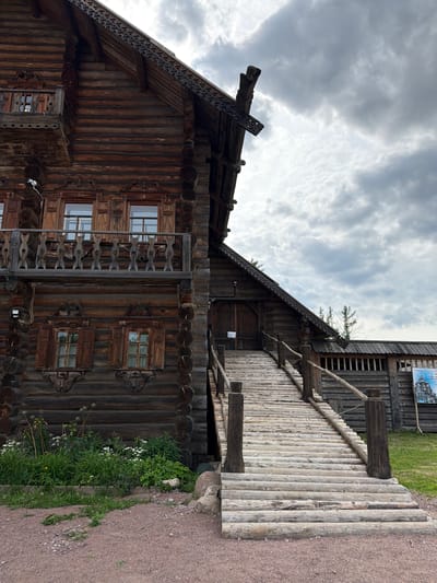 The image shows a traditional wooden building with intricate carvings and a wooden walkway leading up to it. The building appears to be constructed from logs, with several windows featuring decorative wooden shutters. A balcony with carved railings is visible on the side of the building. The walkway is made of many smaller logs laid side-by-side. In the foreground, a patch of greenery with white flowers and tall grass grows alongside a gravel path. The sky is overcast with dramatic, textured clouds. The overall scene suggests an outdoor historical or cultural site, possibly a museum or heritage park. There is a poster on the wall of a structure behind the main building, but the text is not legible. The lighting and cloudy sky suggest it might be daytime, possibly late afternoon or a generally overcast day. The location is identified as Dnp Ryzhiki, Russia.