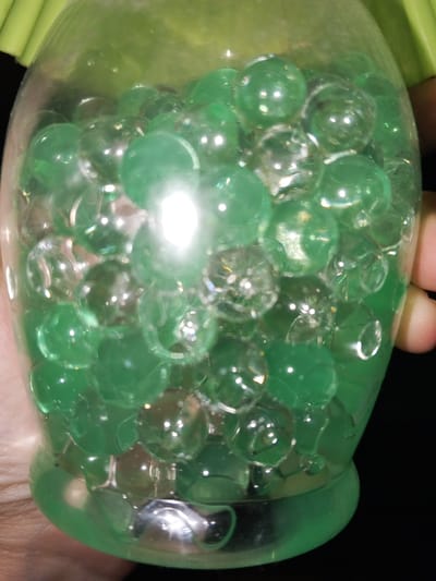 The image displays a close-up view of a clear, translucent container, possibly glass or plastic, held by an individual's hands, with portions of the hands visible on both sides against a dark background. The container is densely packed with numerous spherical objects, primarily green and a lesser quantity of clear ones, consistent with water beads or gel balls. A strong, bright light source creates prominent white reflections on the curved surfaces of multiple spheres. The lower section of the container has a distinct green hue. A blurred green object is partially visible at the upper left edge of the frame. This scene is located in the city of Kabal, Pakistan.