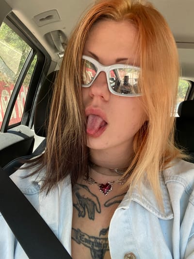 A young person with blonde and brown hair, wearing white-framed glasses and a light blue denim jacket, is shown from a close-up, tilted perspective. The person is sticking their tongue out. The person has tattoos on their neck and collarbone, and a silver necklace with a red pendant. They appear to be in a car, wearing a seatbelt. The reflection in the glasses shows a building and trees outside, possibly in Kruče - Kërruçi, Montenegro.