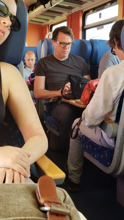 The image depicts an interior view of a train carriage with passengers. In the foreground, the left arm and hand of a person wearing a black top and sunglasses are visible. To the right of this person, a man with glasses and earbuds is seated, looking down. Further back, another man with a bald head and earbuds is also seated. A red bag and a black bag are placed on a tray table in front of the man with glasses. The seats are blue with an orange and blue pattern. The train carriage has orange curtains and windows. The flooring is dark blue. This scene appears to be from a train traveling through or near Horná Streda, Slovakia, given the context.