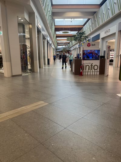 The image captures an interior view of a modern shopping mall, likely located in Limbiate, Italy, given the context. The scene is dominated by a wide, tiled walkway with a light wood-colored inlay in the foreground. The walkway leads into the distance, flanked by shopfronts on either side. On the right side of the image, a "Punto Info" (Information Point) kiosk is visible, featuring a screen displaying animated characters and the BRT logo. A red fire extinguisher stands to the left of the kiosk. Several individuals are present in the mall. In the middle ground, two men, one in a dark uniform with yellow accents and the other in a light blue shirt, appear to be standing near the information point. Further down the mall, other shoppers are seen walking. The mall's architecture includes a high ceiling with natural light filtering through a central skylight structure, and the shop facades are clean and contemporary. The overall atmosphere suggests a typical daytime scene in a retail environment.