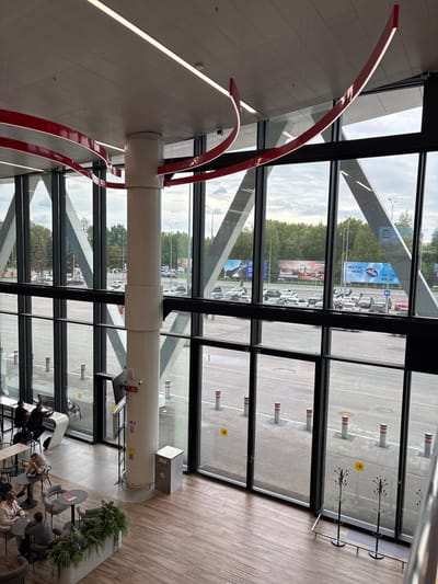 This image depicts an interior view of a modern building with large windows overlooking an outdoor parking lot and a street. The interior features a white cylindrical pillar with red curved decorative elements extending from the ceiling. The floor is made of light brown wooden planks.

Several people are visible inside the building, seated at tables in what appears to be a waiting area or lounge. They are engaged in various activities, such as looking at their phones or talking to each other, suggesting a relaxed or waiting atmosphere.

Outside, the parking lot is filled with numerous cars. Large billboards are visible in the distance, some displaying text. The weather appears to be overcast, with a cloudy sky. The overall scene suggests a public space, possibly an airport or a large commercial building, given the scale of the windows and the presence of parking and billboards. The digital clock on the pillar indicates the time is 17:26. Some text on the billboards is partially visible, including "ЧИСТОЕ НЕБО" (CLEAN SKY) and "ЭРОДЫ КОЛОКОЛА" (EROS OF THE BELL). The location context is Старое Мартьяново, Russia.