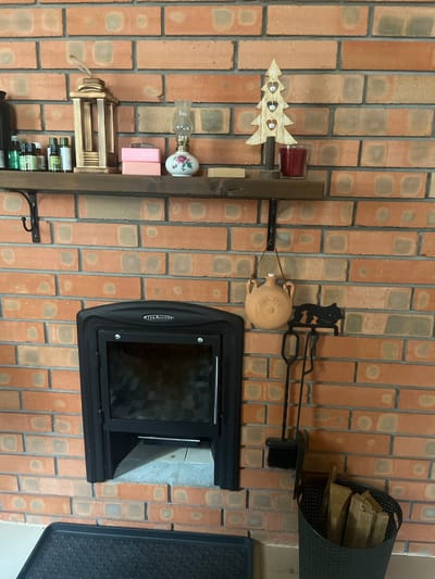 The image depicts an indoor scene, likely a living room or a cozy nook, against a rustic brick wall. The focal point is a black wood-burning stove with a glass door, set into a recess in the wall. Above the stove, a wooden shelf displays a variety of decorative items. These include a wooden lantern, several bottles of what appear to be essential oils, two pink soap bars, a small oil lamp with a floral design, a wooden Christmas tree ornament, and a red candle in a glass holder. To the left of the stove, a black metal hearth-cleaning set, consisting of a brush, shovel, and poker, hangs on the wall. Below the shelf and to the right of the stove, a decorative terracotta jug with a leather handle is also suspended. A black basket filled with firewood is situated next to the stove. The overall aesthetic is warm and inviting, with a blend of rustic and decorative elements. The image appears to be taken in Староживотинное, Russia.