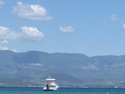 The image depicts a serene coastal scene in Lichada, Greece. In the foreground, the clear turquoise waters of the sea are visible, with a small white boat with red accents anchored in the middle. The boat has multiple decks and appears to be a passenger vessel. A single red buoy floats in the water to the left of the boat. In the background, a range of lush green hills and mountains forms a picturesque backdrop against a bright blue sky dotted with white, fluffy clouds. A thin strip of coastline with scattered buildings is visible at the base of the mountains. The overall atmosphere is tranquil and idyllic, characteristic of a Mediterranean summer day.