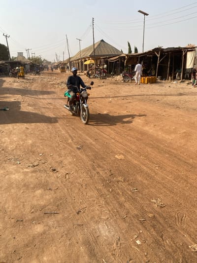 A dusty, sunlit street scene features a man riding a motorbike. He's traveling on the left side of the road which is made of light brown dirt. The road shows many tire tracks. A row of simple shops lines the left side of the road, with people walking around. Power lines and poles run alongside the buildings. The scene has a dry, rural atmosphere.