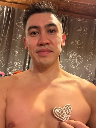 A man poses shirtless with a heart-shaped cookie resting on his chest. He has dark hair, fair skin, and is smiling slightly. The background features a curtain with a gray rose pattern. A glimpse of what appears to be a wooden structure or beam is visible in the top left corner. The lighting casts shadows on his body, highlighting his chest and shoulders. The image appears to be taken from a low angle.
