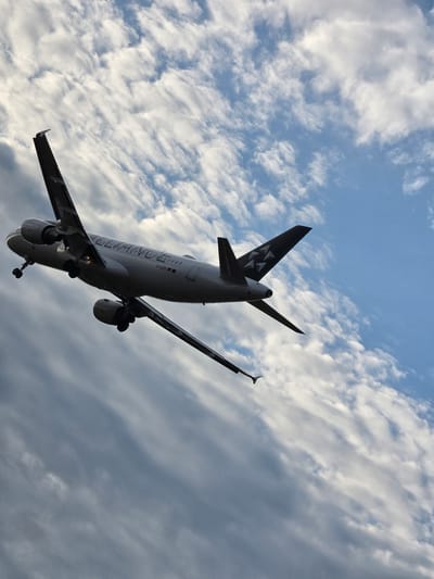 An airplane is captured in flight, angled upwards against a sky filled with dynamic clouds. The aircraft, appearing to be a passenger jet, has its landing gear extended, suggesting it is either approaching or departing from an airport. The fuselage is predominantly white with prominent branding, including the word "ALLIANZ" and a registration number "D-AIZM" visible. The tail fin is dark, adorned with a stylized triangular logo. The engines are mounted under the wings. The background consists of a mix of bright white and darker gray clouds against a blue sky. This image was taken near Kelsterbach, Germany, a location known for its proximity to Frankfurt Airport.