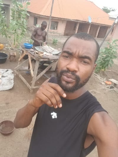 The image shows a man, dressed in a black sleeveless shirt, taking a selfie in front of another man who is butchering a rabbit on a wooden table. The scene is set outside in an open area, possibly a courtyard, with a building in the background. The building appears to be a residential structure, with a tiled roof and windows. There are plants in the foreground and some small, scattered objects on the ground. The man taking the selfie has a beard and is looking directly at the camera. The man butchering the rabbit is facing away from the camera and is wearing a black shirt. The setting is Pati Ridge, Nigeria, a residential area located at the coordinates provided.