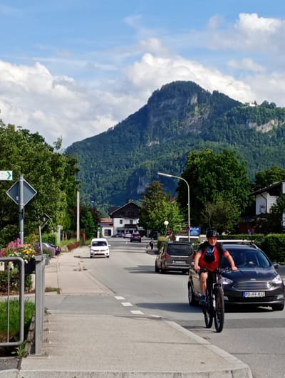 The image depicts a street scene in Brannenburg, Germany, on a sunny day with scattered clouds. A man is riding a bicycle towards the camera, wearing a helmet and athletic clothing. He is on the right side of a two-lane road, in front of a dark-colored car with a license plate that reads "RO FF 812". The road is lined with trees and buildings. On the left side of the road, there is a sidewalk with a flower planter and a street sign. In the background, a large, tree-covered mountain dominates the skyline, with a building visible near its peak. The overall scene suggests a typical village or town environment in a mountainous region.