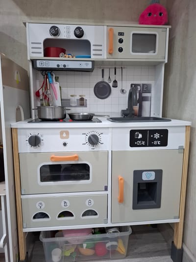 A child's wooden toy kitchen set is positioned against a textured, light-colored wall. The kitchen set is white and light grey with wooden accents. On the top shelf, a pink plush toy with large googly eyes sits on the right. The kitchen features a microwave with a digital display showing "02:30", an oven with knobs, a stovetop with two burners, a sink with a faucet, and a refrigerator with a digital display showing temperatures of +05°C and -18°C. Below the oven, there are three compartments with icons for recycling: a glass bottle, an apple core, and a water bottle. A clear plastic bin filled with various toys, including colorful fruits and a pink plush toy, is placed beneath the kitchen set. The floor is covered with light grey wooden planks. The environment appears to be indoors, likely a playroom or child's room. The overall scene is well-lit and shows a clean, organized toy setup. There is no indication of Malaya Tsarevschina, Russia.