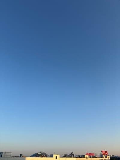 The image shows a clear blue sky above a low-rise urban landscape. The horizon is marked by buildings of varying heights, with some structures featuring signage. One prominent building has a domed roof. The lighting suggests it is either sunrise or sunset, casting long shadows. There are no visible people in the image. The scene is likely part of the city of Sovkhoza imeni Lenina, Russia, given the context.