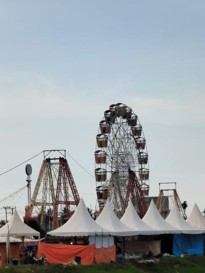 This image, taken in Blangbitra, Indonesia, shows a fairground under an overcast sky. A Ferris wheel and other carnival rides are visible behind a row of white, conical tents. An orange and a blue cloth are draped over some of the tent openings. People and a motorcycle can be seen near the tents. Grass covers the ground in front of the tents. Power lines run overhead.