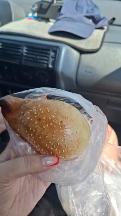 A person's hand, with red and white french tip fingernails, holds a bread roll inside a clear plastic bag. The bread roll is golden brown, sprinkled with sesame seeds, and appears to have a slightly darker, toasted end. The background suggests the interior of a car, with a black dashboard, air vents, and a blue baseball cap resting on a light-colored dashboard mat. A small toy is also visible on the mat. The overall scene is illuminated by natural light, indicating it's daytime. The location is identified as Қасымбек датқа, Kazakhstan.