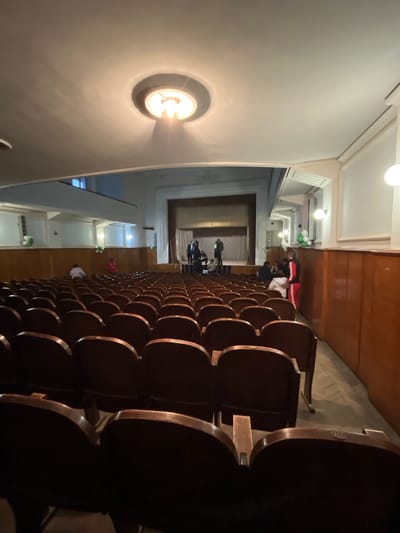 The image shows the interior of a theater or auditorium, oriented vertically. Rows of empty dark brown or black seats fill the right side of the frame, suggesting a large seating capacity. People are visible in the background toward the stage area, some standing and some sitting. The stage itself appears to be covered by a curtain. The left side of the image shows a wall with a circular light fixture illuminated, emitting a warm glow. The environment appears to be somewhat old-fashioned, with classic architectural details. It is not possible to determine the location within Voiniagovo, Bulgaria from this image.