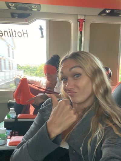A medium shot captures a blonde woman making a playful pout and giving a thumbs-up sign to the camera, her right hand positioned near her chin. She is seated in what appears to be a vehicle, possibly an ambulance or bus, with an orange interior. In the background, another person is visible, wearing a blindfold and red socks, with their legs crossed. The vehicle's window offers a view of buildings and trees. The text "Hotline" is visible in reverse on the window. The lighting suggests it is daytime, with sunlight streaming in.