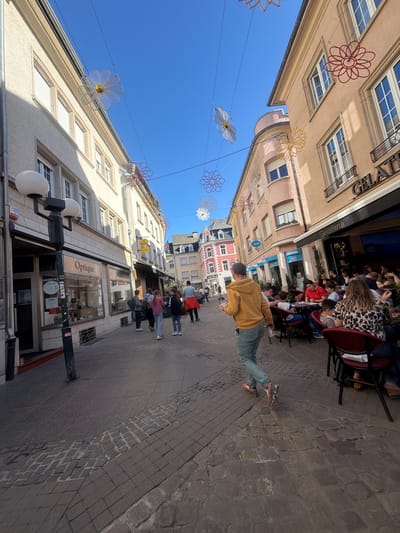 The image depicts a street scene in Echternach, Luxembourg, characterized by a blend of commercial and residential buildings under a clear, blue sky. The viewpoint is along a cobblestone street that curves slightly, lined on both sides by buildings of varying heights and architectural styles. Decorative flower-like ornaments are suspended across the street, adding a festive or seasonal touch.

On the right side of the street, people are seated at an outdoor cafe, suggesting a leisurely atmosphere. Some are wearing masks. Further down the street, small groups of people are seen walking. On the left, commercial storefronts are visible. One storefront is identified with a sign "Optique", indicating an optician or eyewear shop. A street lamp is positioned near this storefront.

A person is walking away from the camera wearing a yellow hoodie and green trousers. They are walking on the cobblestone street.