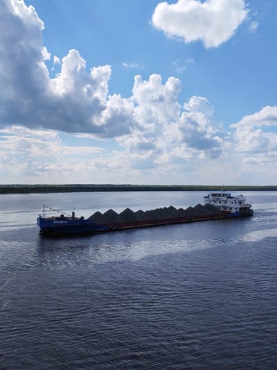 The image depicts a barge laden with what appears to be dark granular material, possibly coal or ore, traveling on a wide body of water. The barge is a blue and white, multi-sectioned vessel, with the name "OKSKII-67" visible on the front. The background shows a distant line of trees along the horizon. The sky is partly cloudy with patches of blue. Pruzhinino is not visibly identifiable within the frame, and its presence is purely circumstantial.