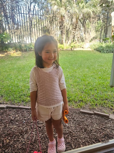 A young girl stands in a grassy yard bordered by wood chips and a low retaining wall. She wears a white shirt, light-colored knitted vest, white shorts, pink socks, and pink Crocs. She holds orange binoculars in one hand and red jump ropes in the other. The background includes green grass, foliage, trees, and a fence. The location is La Molina, Peru, based on the foliage and architectural features.