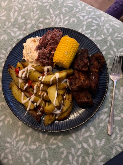 This image depicts a prepared meal served on a dark blue, ribbed ceramic plate, located in the city of New Forest, United Kingdom. The plate contains distinct portions of food: a pile of roasted potato wedges seasoned with green herbs and red spices, drizzled with a white sauce; a serving of creamy coleslaw; a portion of shredded, reddish-purple braised cabbage; a bright yellow half-ear of corn on the cob; and several pieces of dark, glazed barbecued ribs. A silver-colored metal fork, featuring a textured handle, is positioned to the right of the plate. Both the plate and fork rest upon a soft, light green textile surface adorned with a repeating pattern of lighter botanical motifs. No individuals or active interactions are present in the scene.